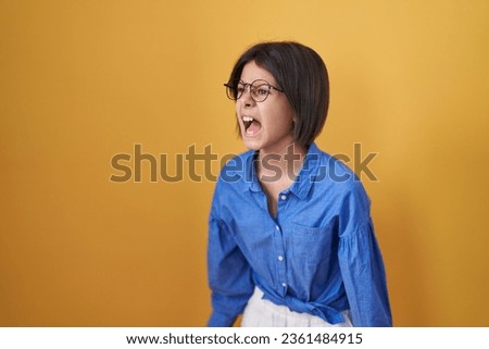 Young girl standing over yellow background angry and mad screaming frustrated and furious, shouting with anger. rage and aggressive concept. 