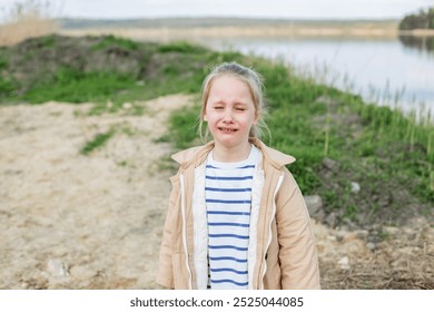 Young girl stands by riverbank, visibly upset and crying, with natural outdoor backdrop of grass and water. - Powered by Shutterstock