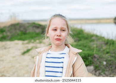 Young girl stands by riverbank, visibly upset and crying, with natural outdoor backdrop of grass and water. - Powered by Shutterstock