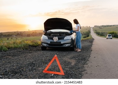 A young girl stands near a broken car in the middle of the highway during sunset and tries to repair it. Troubleshooting the problem. Waiting for help. Car service. Car breakdown on road.