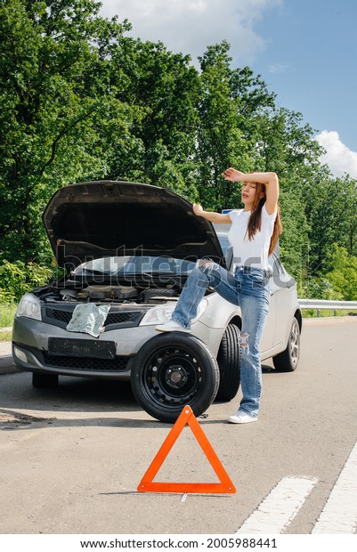 A young girl\
stands near a broken car in the middle of the highway and tries to\
change a broken wheel on a hot sunny day. Failure and breakdown of\
the car. Waiting for help.