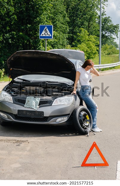 A young girl stands near a broken car with a broken\
wheel in the middle of the highway and is frustrated waiting for\
help on a hot day. Breakdown and breakdown of the car. Waiting for\
help.