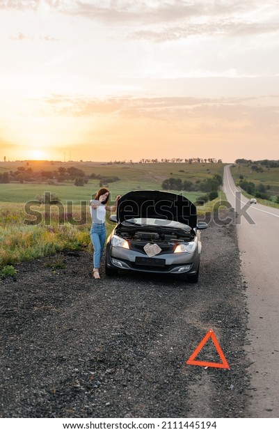 A young girl stands near a broken car in the middle\
of the highway during sunset and tries to call for help on the\
phone and start the car. Waiting for help. Car service. Car\
breakdown on the road.