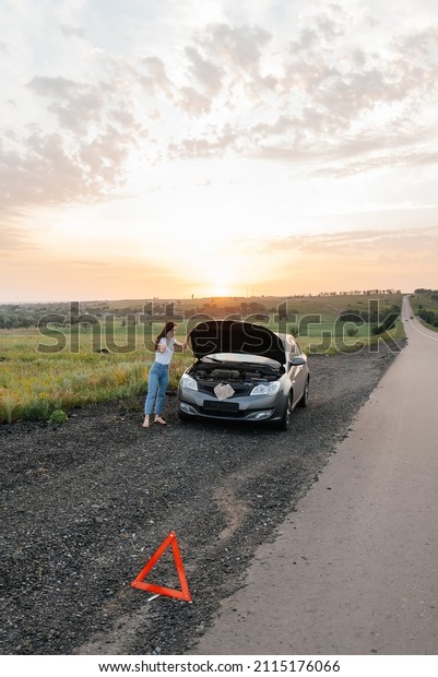 A young girl stands near a broken car in the middle\
of the highway during sunset and tries to call for help on the\
phone and start the car. Waiting for help. Car service. Car\
breakdown on the road.