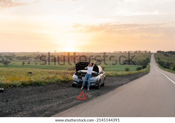 A young girl stands near a broken car in the middle
of the highway during sunset and tries to call for help on the
phone and start the car. Waiting for help. Car service. Car
breakdown on the road.