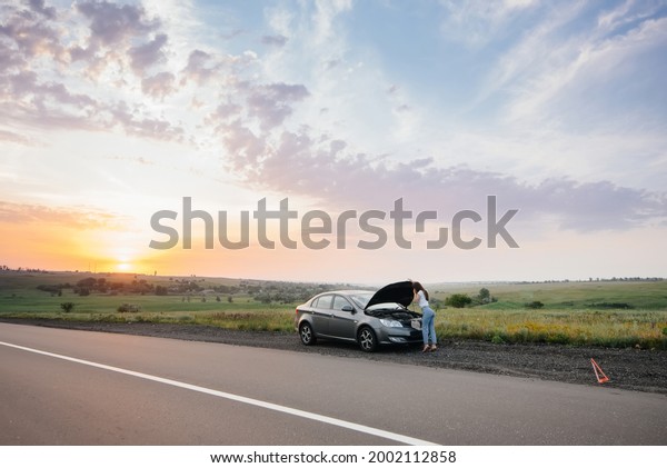A\
young girl stands near a broken-down car in the middle of the\
highway during sunset and tries to call for help on the phone.\
Breakdown and repair of the car. Waiting for\
help.