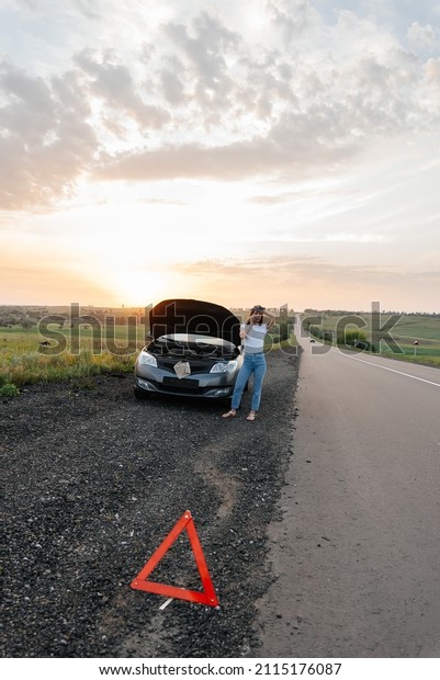 A\
young girl stands near a broken-down car in the middle of the\
highway during sunset and tries to call for help on the phone.\
Waiting for help. Car service. Car breakdown on\
road.