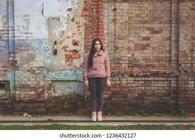 Young Girl Stands On The Background Of An Old Dirty Dilapidated Wall. She's Wearing A Pink Jacket, Gray Jeans, Pink Ugg Boots. Her Hair Is Long And Black.