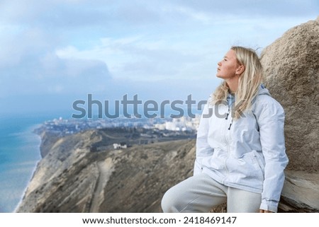 A young girl stands on a high mountain and looks at the sea and a beautiful landscape on a sunny day. Cute smiling blonde with flowing hair wearing a white jacket. Active lifestyle and recreation.