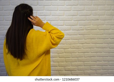 Young girl with straight dark hair in a yellow sweater against a white brick wall. Hands in hair - side view