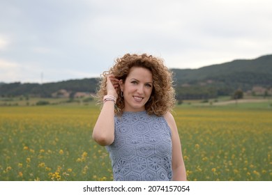 Young Girl Stroking Her Curly Blond Hair In The Meadow, Wearing A Blue Sleeveless Sweater