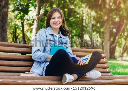 Young girl student sits on a bench in the park and holds books, notebooks and mobile phone. Girl teaches lessons in the park.