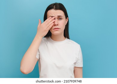 Young girl suffering from eyes pain and feeling something in eye, posing on blue wall. Cause of pain include contact lens problem, conjunctivitis, foreign object, dry eye syndrome or allergy