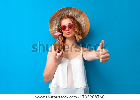 young girl in summer clothes drinks a coconut cocktail and shows like on a blue isolated background, woman tourist in a summer resort