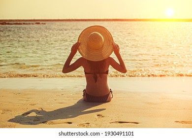 Young Girl At Sunset On The Beach Sits With Her Back To The Sea In A Hat