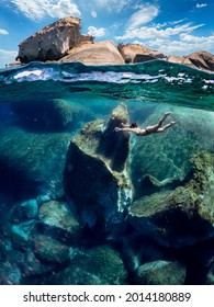 Young Girl Swimming Underwater In Deep Water. Woman In Half Underwater Effect While She Swims Surrounded By Huge Rocks. Villasimius, Sardinia.