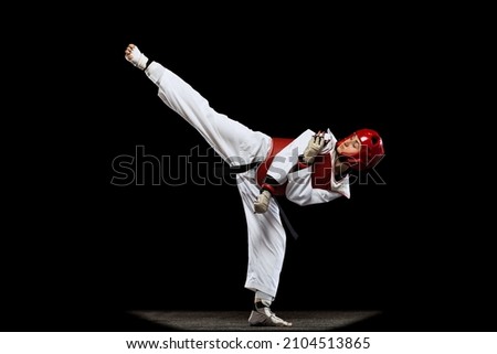 Young girl, taekwondo practitioner strikes forcibly with the foot isolated over dark background. Concept of sport, education, skills, workout, health. Sportsman wearing white dobok