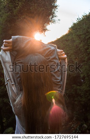 
young girl taking off a gray sweater in a natural maze with a very orange sunset