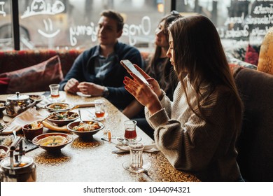 Young Girl Is Taking Pictures Of Food. A Young Company Of People Is Smoking A Hookah And Communicating In An Oriental Restaurant. Lebanon Cuisine Served In Restaurant.  Traditional Meze Lunch