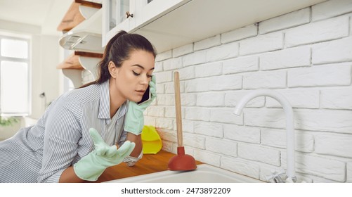 Young girl talking by the phone advising how to unclog a sink drain. Girl trying to clean sink using plunger standing in the kitchen at home. Housekeeping and household work concept.