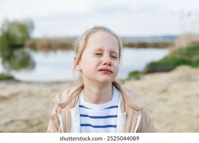 Young girl with tears in eyes stands outdoors near calm lake, visibly upset and emotional on peaceful day. - Powered by Shutterstock