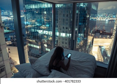 Young girl or teenager with long hair girl checking her mobile phone or chatting with someone at night in her bed with breathtaking view over night city. Hong Kong, China. - Shutterstock ID 401782333