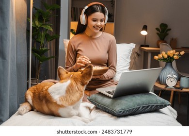 Young girl, teenager sitting on her bed with her dog, wearing headphones and watching a movie on her laptop while eating popcorn from a wooden bowl - Powered by Shutterstock