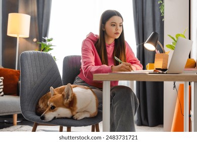 Young girl, teenager studying at home, focused on her laptop and jotting down notes, with her corgi dog resting nearby. Natural light creates a cozy atmosphere for e-learning - Powered by Shutterstock