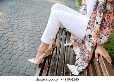 Young Girl In Trendy Colorful Cardigan Sitting On The Wooden Bench In The Park. Woman In A Pink Cape In High Heels Leather Shoes.