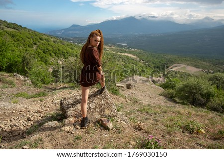 A young girl of unconventional appearance with red African braids in full growth in a brown t-shirt put her foot on a stone, against the blue sky and mountains, under the scorching rays of the sun