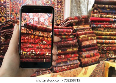  Young girl using smart phone taking photo of handmade persian carpet in the grand bazaar of Isfahan, Iran. Vintage carpets or rugs are very good value for money purchase.