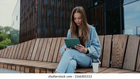 Young Girl Using Tablet Computer Outdoors In The Street While Sitting On The Bench. Pretty Female Tourist Strolling And Looking At Screen, Browsing Internet, Chatting.