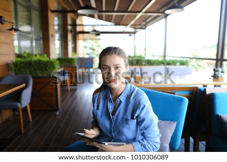 Young girl using tablet at restaurant and sitting on blue sofa. Beautiful girl wears in casual and has pony tail. Concept of wireless local area network at catering establishment and browsing.