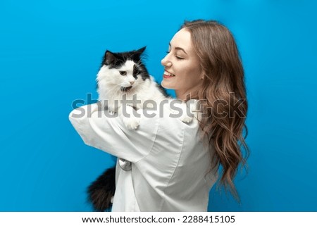 young girl veterinarian in uniform holds black and white cat on blue isolated background, doctor in medical coat with stethoscope looks smart and loves animals