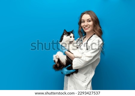 young girl veterinarian in uniform holds black and white cat on blue isolated background, doctor in medical coat with stethoscope looks smart and loves animals
