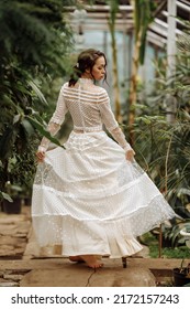 A Young Girl In A Vintage White Dress And A Hat On Her Back Stands Sideways In A Tropical Greenhouse.