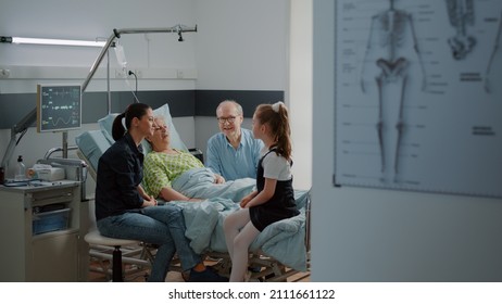 Young girl visiting aged woman in hospital ward bed with mother and old man. Child talking to sick grandma at family visit intensive care room, giving support to hospitalized patient with disease.