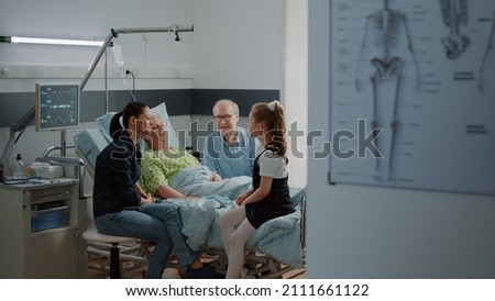 Young girl visiting aged woman in hospital ward bed with mother and old man. Child talking to sick grandma at family visit intensive care room, giving support to hospitalized patient with disease.