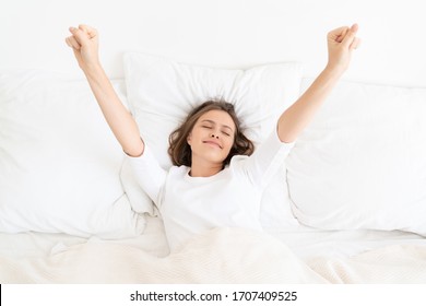 Young Girl Waking Up In Morning In White Bed And Pyjama Top, Leaving Eyes Closed But Stretching Arms Up To Get Ready For Active Day, Showing Sleepy Smile