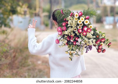 A Young Girl Walks With A Huge Bunch Of Flowers Behind Her Back And Shows The Victory Sign With Her Hand