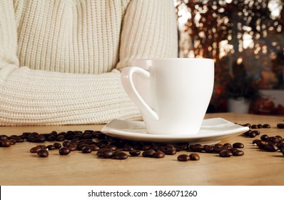 Young Girl In Warm, Cosy Knitted Autumn Sweater, Leaning On A Kitchen Counter, Next To A Cup Of Coffee. Fresh Unground Coffee Beans Scattered On A Wooden Table, With Leaves Behind The Window.