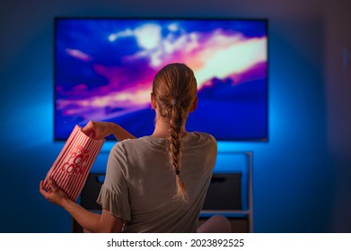 A young girl is watching her favorite movie on TV. She is holding a pack of popcorn. The girl is dressed in home clothes. There is a blue neon light in the room. Rest. Hobby. Day off. - Shutterstock ID 2023892525