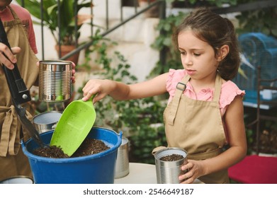 Young girl wearing an apron, gardening and filling recycled tin cans with soil. Engaging in an outdoor planting activity, she demonstrates environmental awareness by using sustainable materials. - Powered by Shutterstock