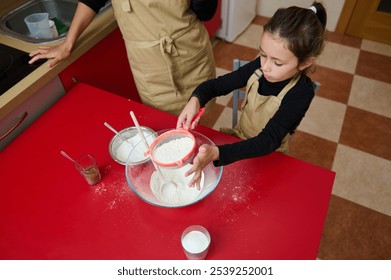 A young girl wearing an apron is sifting flour into a bowl on a red kitchen table. An adult supervises in the background. The scene captures a warm and educational baking activity. - Powered by Shutterstock