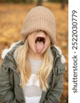 Young girl wearing a beige knit hat pulled down over her eyes, sticking out her tongue in a playful and silly gesture, enjoying the autumn season outdoors with colorful leaves around.
