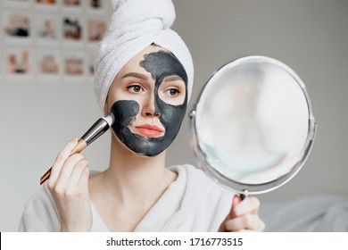 A young girl in a white bathrobe with a towel on her head holds a mirror in her hands and applies a natural black mask with activated carbon for problem skin, quarantine isolation, home spa treatments