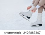 young girl in white and beige winter clothes lacing up skates before skating in ice rink in winter cloudy snowy day, close-up view of skates, copy space