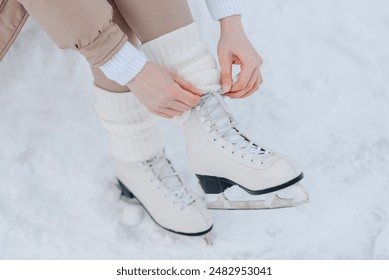 young girl in white and beige winter clothes lacing up skates before skating in ice rink in winter cloudy snowy day, close-up view of skates