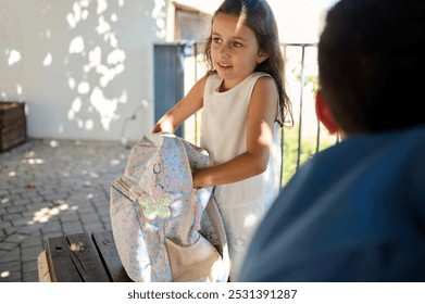 A young girl in a white dress organizes her backpack on a sunny day, reflecting excitement and readiness for school. - Powered by Shutterstock