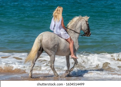 Young girl in white dress rides a white horse on the beach